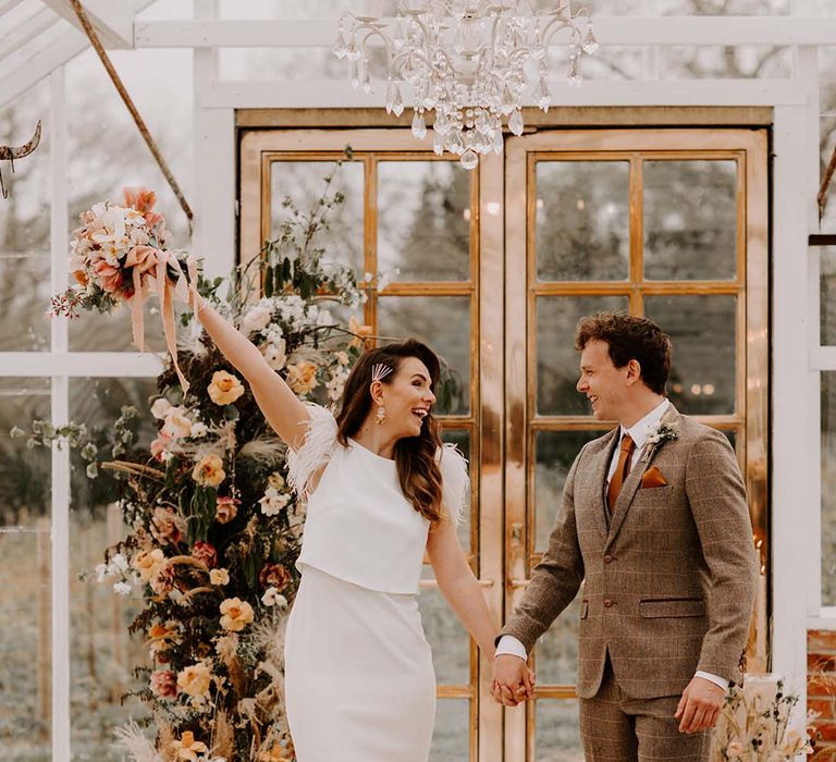 Bride in a fitted wedding dress with puddle train and feather sleeves holding her grooms hand in a brown check suit waving her bouquet in the air as they stand at the altar in The Gooseberry House