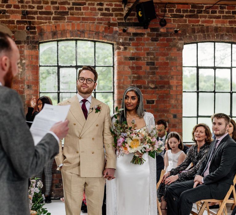 Groom in double breasted linen suit and burgundy tie holds hands with bride in satin Halfpenny London wedding dress and tasselled bridal cape holding multicoloured bridal bouquet during wedding ceremony at Loft Studios London