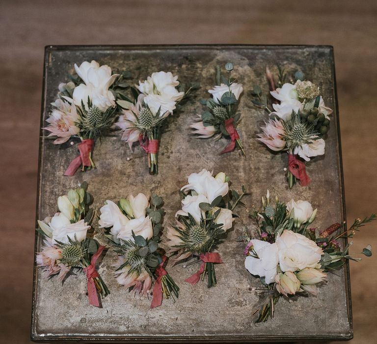 Eight protea, rose and thistle buttonholes tied with red ribbon at Tythe Barn wedding with barn wedding flowers