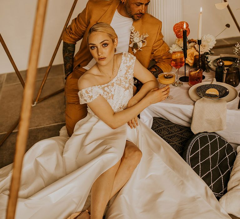 Bride in a Jesus Peiro wedding dress with lace off the shoulder bodice sitting at the relaxed reception with her groom in a mustard suit sipping cocktails 