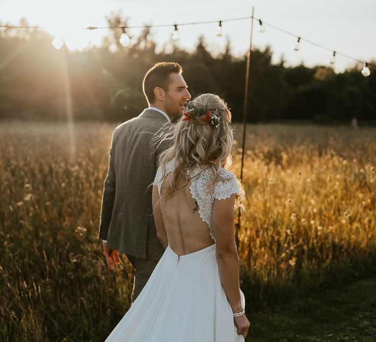 Bride in lace cap sleeved open back wedding dress with train and flowers in her hair walks with groom through field during late summer wedding at Wellington Wood Norfolk