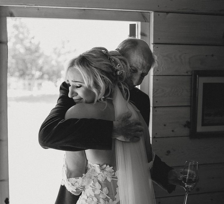 Bride in off the shoulder Enzoani wedding dress and veil smiles as she bugs father in suit holding glass of sparkling wine before tipi wedding at Inkersall Grange Farm