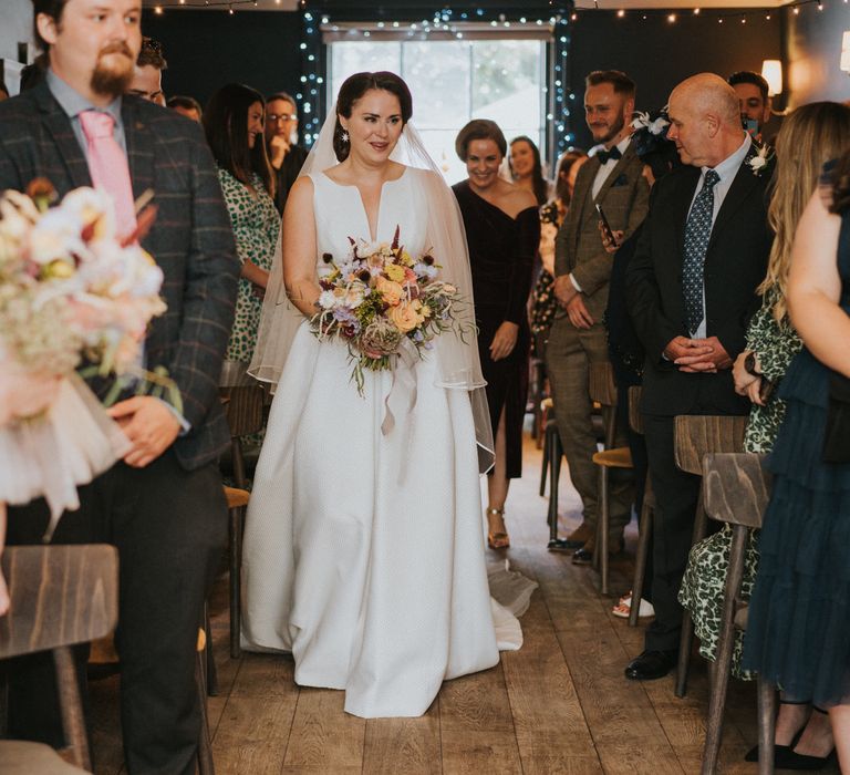 Bride walks down the aisle in Jesus Peiro wedding dress carrying pastel flower bouquet