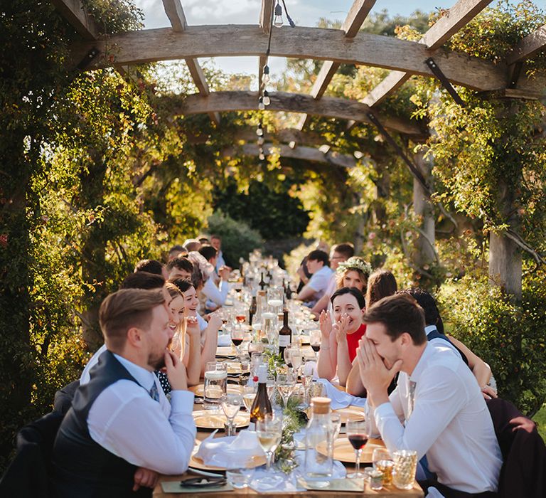 Wedding guests eat outdoors on banquet table at Euridge Manor surrounded by green foliage under pergola