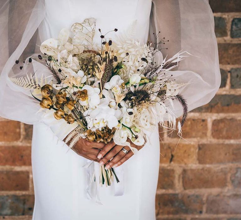 Bride in a sheer long sleeve wedding dress holding a dried grass and fresh flower wedding bouquet with white orchids 