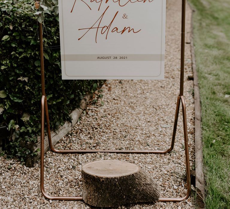 Copper frame wedding sign reading 'Welcome to the wedding of Kathleen and Adam August 28 2021' with foliage decor