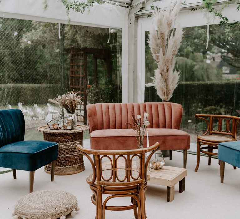 Pink velvet sofa and two blue velvet occasion chairs with various wooden furniture in corner of marquee at wedding reception with garlands and pampas grass for garden wedding