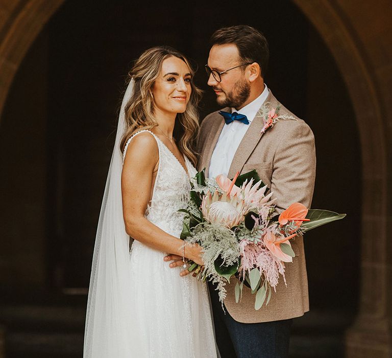 Bride & groom stand outside church whilst bride holds pastel floral bouquet and groom wears pale brown jacket