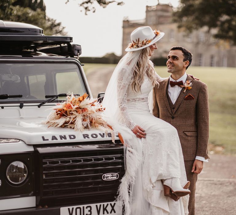 Bride in lace boho two piece wedding dress, bridal hat and veil sits on hood of Land Rover with arm around groom in three piece brown tweed suit in the grounds at Kinmount House in Scotland 