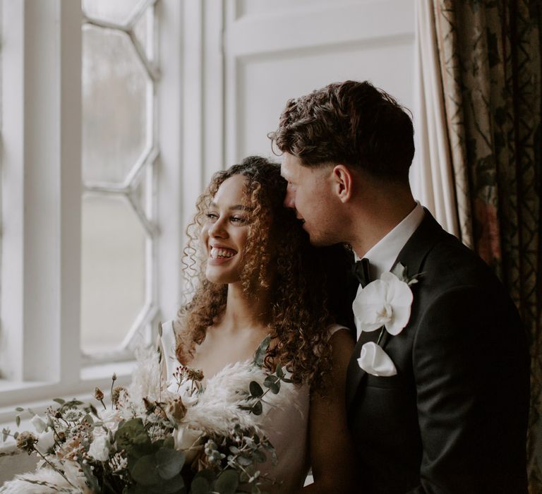 Bride & groom stand beside one another at window on their wedding day