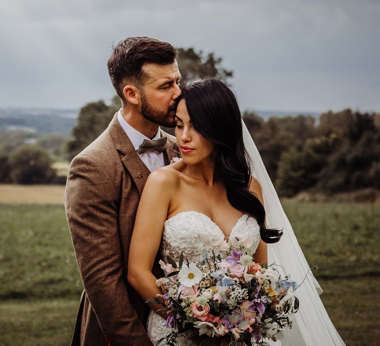 Bride in lace wedding dress with sweetheart neckline and veil holding summer pastel wedding bouquet stands in field with groom in brown tweed suit in the Cotswolds