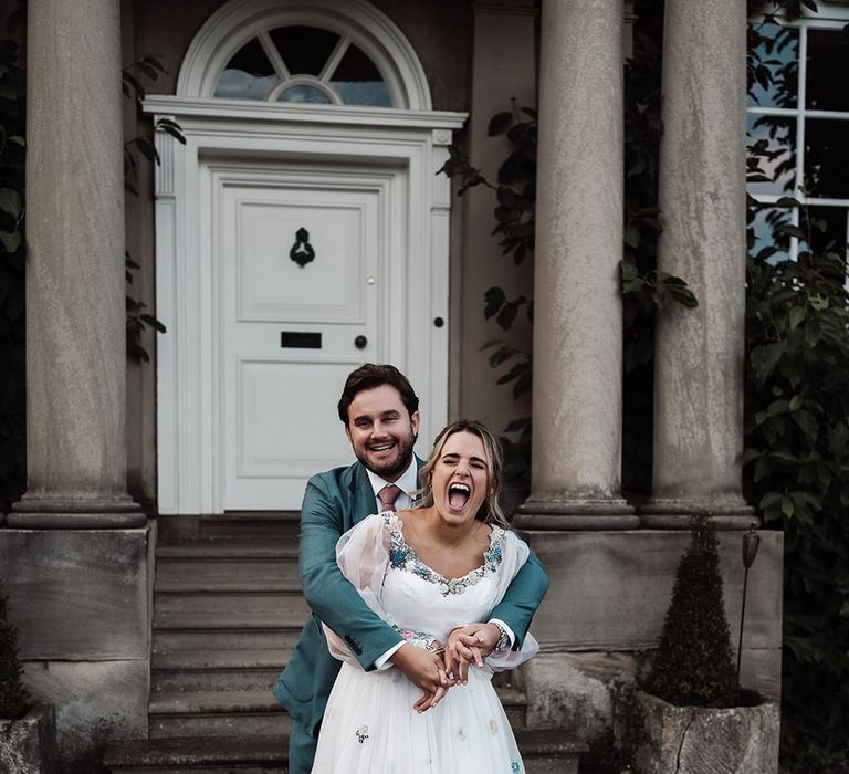 Laughing bride in long sleeve wedding dress with floral embroidery is hugged by groom in blue suit outside the front of Iscoyd Park