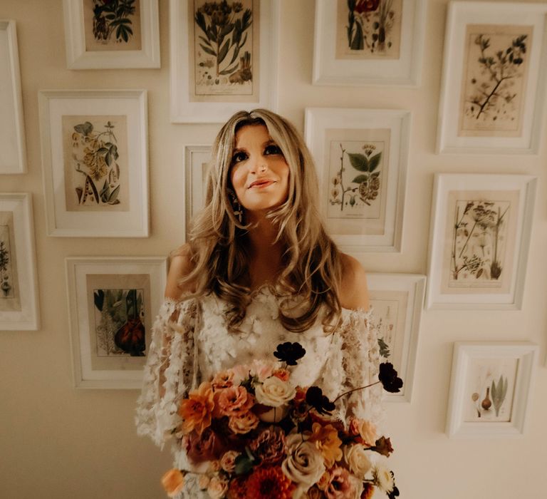 Bride in off the shoulder 3D flower applique wedding dress holding mixed autumnal bridal bouquet stands in front of wall of framed wildflower prints