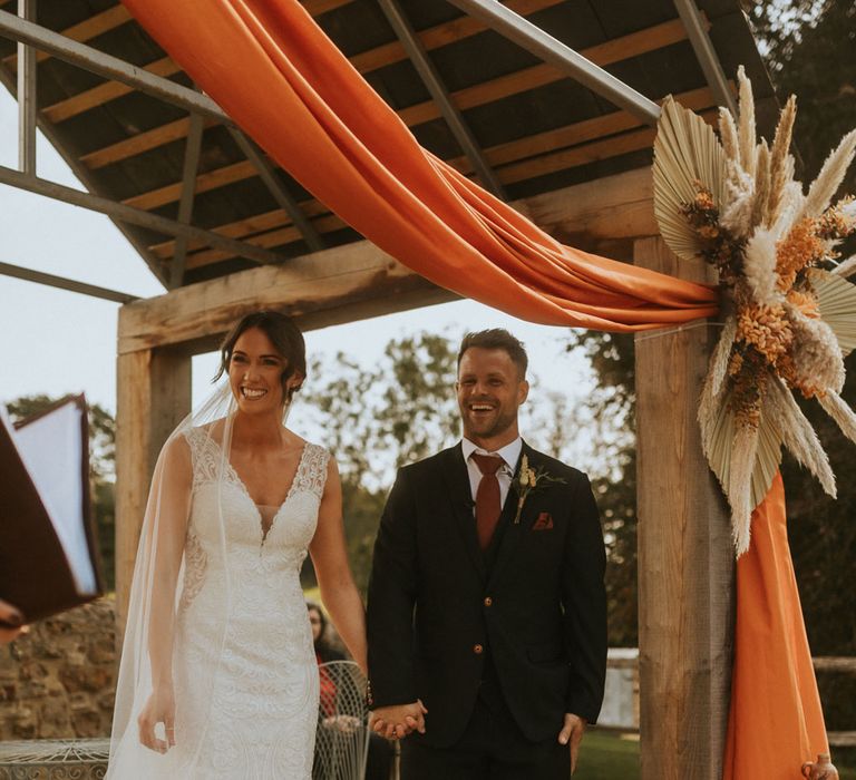 Bride in lace wedding dress and veil stands under orange draping with groom in dark suit, burgundy tie and brown shoes for outdoor wedding ceremony in Dorset