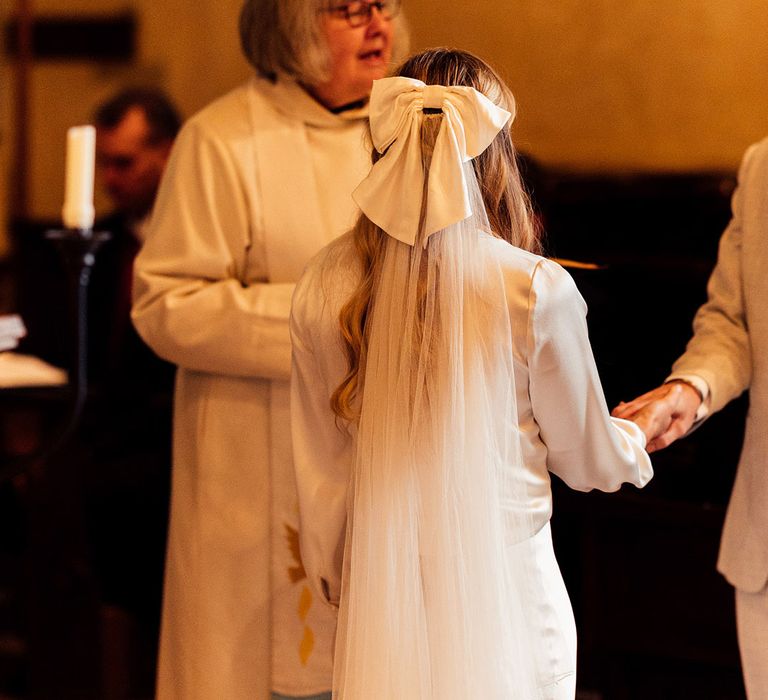 Bride with white bridal hair bow and veil stands with groom in light grey suit, open white shirt and floral buttonhole during church wedding ceremony