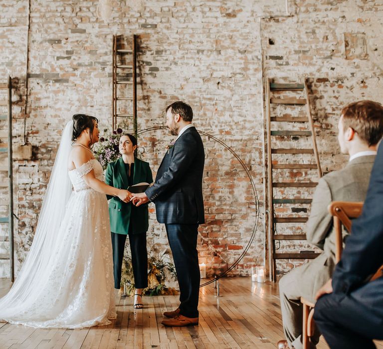 Bride in off the shoulder wedding dress, veil and pearl necklace stands holding hands with groom in blue suit and brown shoes during non-denominational wedding ceremony