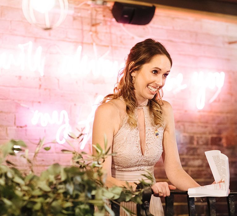 Bridesmaid in soft muted pink dress reads out speech in front of pink neon sign at wedding venue in Essex