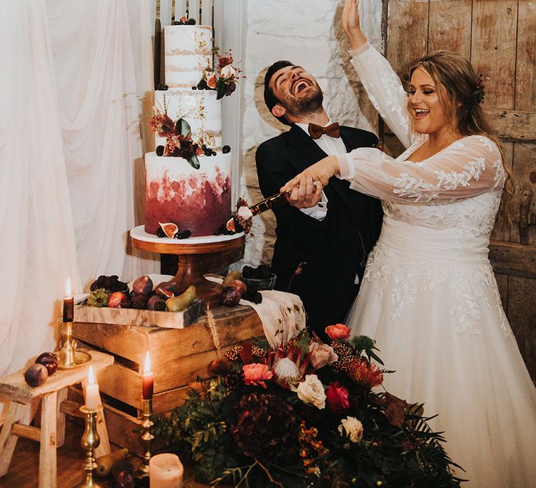 curvy bride in a long sleeve wedding dress cutting the white, burgundy and gold wedding cake on a rustic crate cake stand 