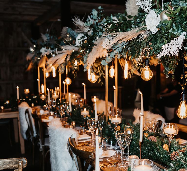 White fur throws on backs of chairs with pinecone runner, white candles, gold-rimmed glasses and exposed lightbulbs