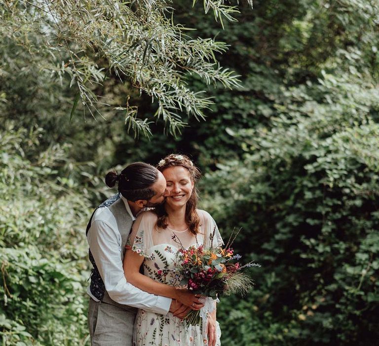 Groom in grey trousers and waistocat kissesthe bride on the cheek who wears a floral embroidered wedding dress with colourful bouquet 