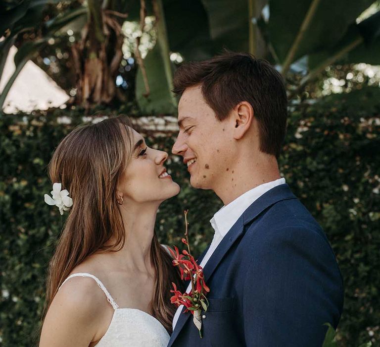 Groom in blue suit and red tropical flower buttonhole with bride in fitted lace beach wedding dress