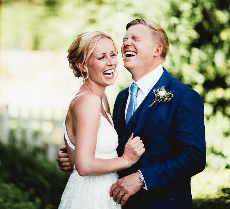 Bride in open back with criss cross straps on boho lace wedding dress laughs with groom in three piece suit