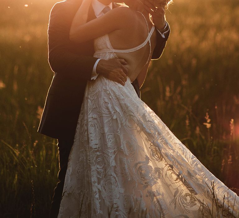 Bride with sparkly hair accessory and boho lace wedding dress with open back kisses groom in blue suit at golden hour