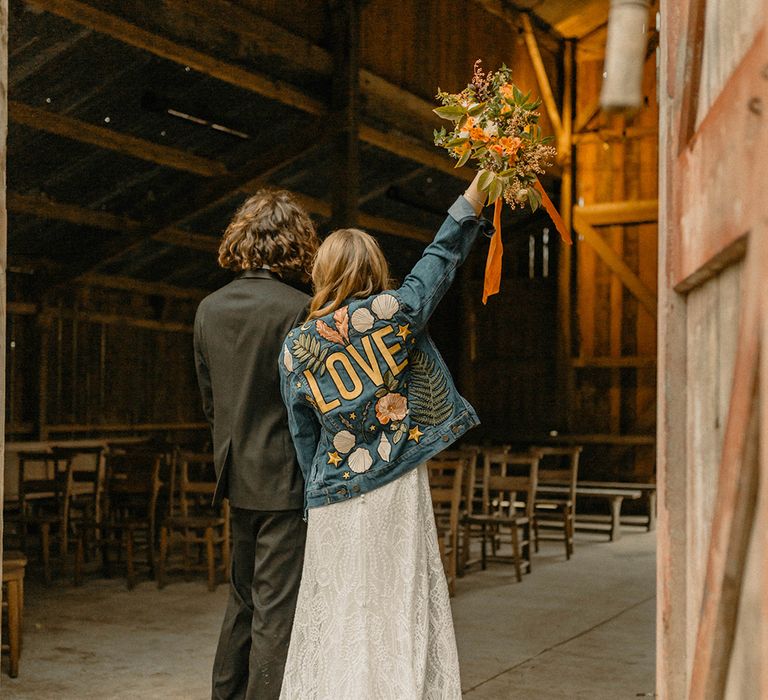 Bride in embroidered denim jacket raising autumnal bouquet with orange ribbon holding hands with groom in black tie