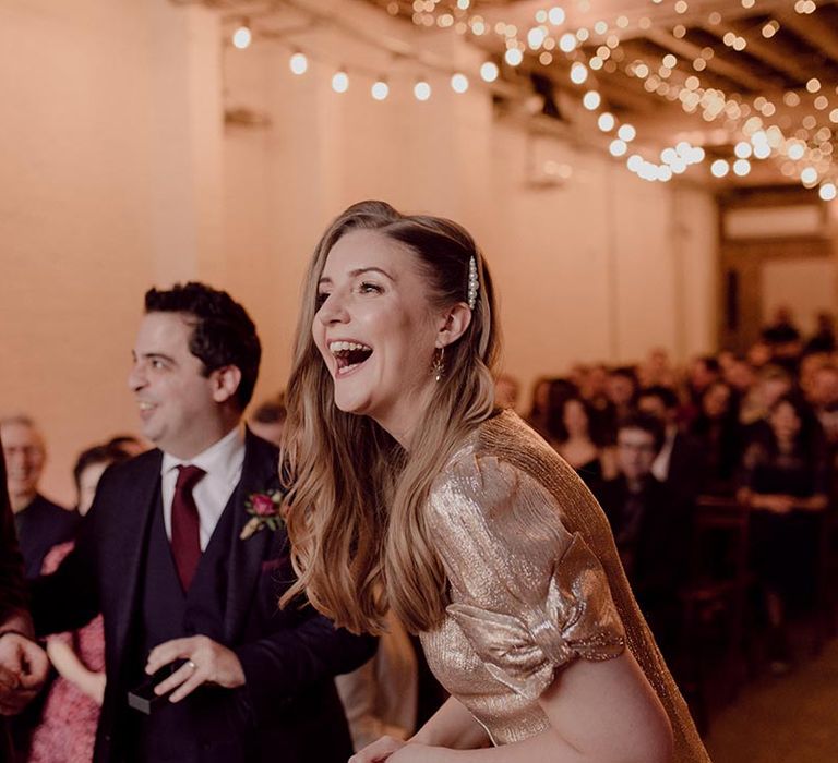 Bride in sparkly gold wedding dress with bow sleeves and pearl hair clip smiles standing at the aisle with the groom in a velvet red suit jacket