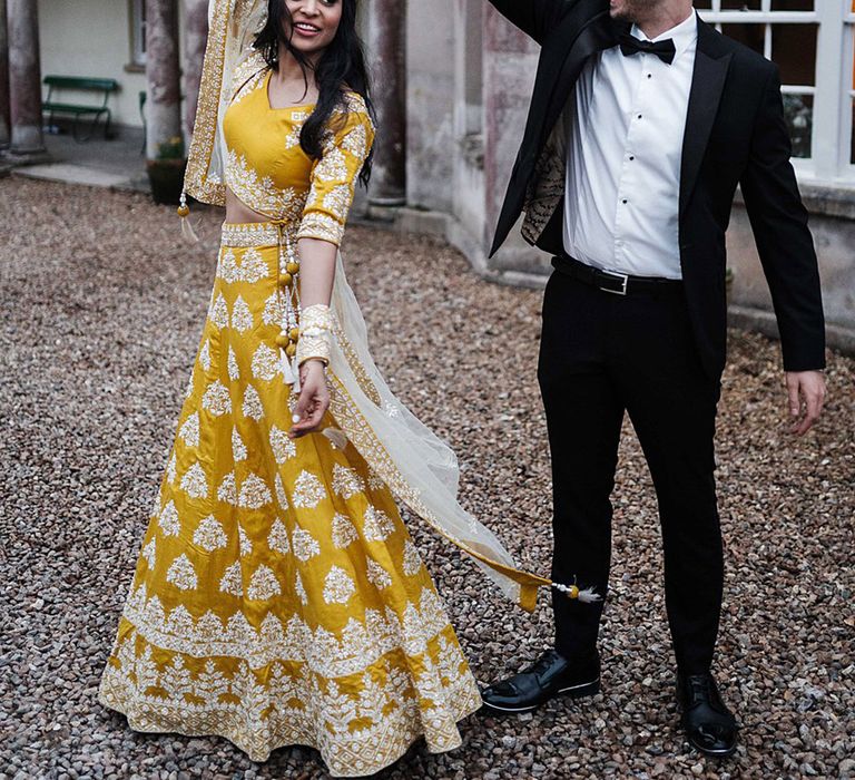 Groom in black tie spins the bride around wearing yellow Lengha outside Pennard House wedding venue 