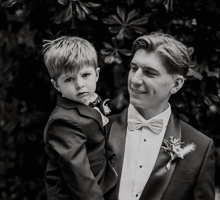 Groom in black tie with pale blue bow tie holds son in black tie for wedding 