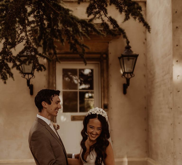 Groom in neutral wedding suit holds the bride around her waist while she wears an Alexandra Grecco wedding dress and flower bridal headband