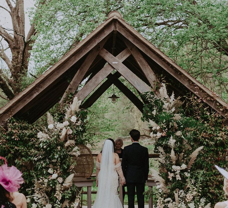 Bride and groom stand holding hands at the altar for their outdoor wedding at Millbridge Court with dried grass and flower column decor 