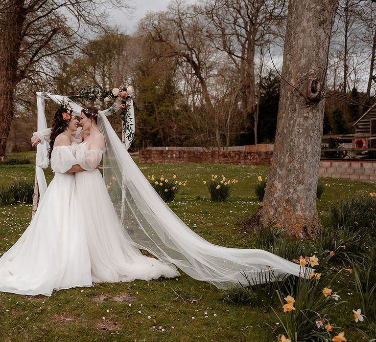 Two brides in ethereal wedding dresses at outdoor wedding ceremony at Mapledurham wedding venue in Oxfordshire with wooden frame decorated with drapes and flowers 