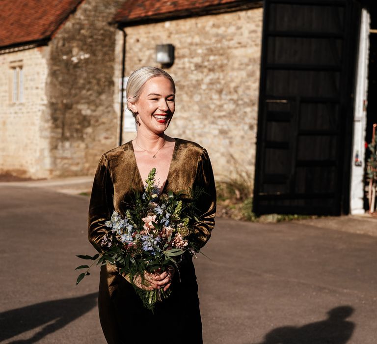 Bride with slicked back blonde hair and red lipstick wears olive green velvet wedding dress from Reiss with V-neck front whilst holding pastel bridal bouquet 