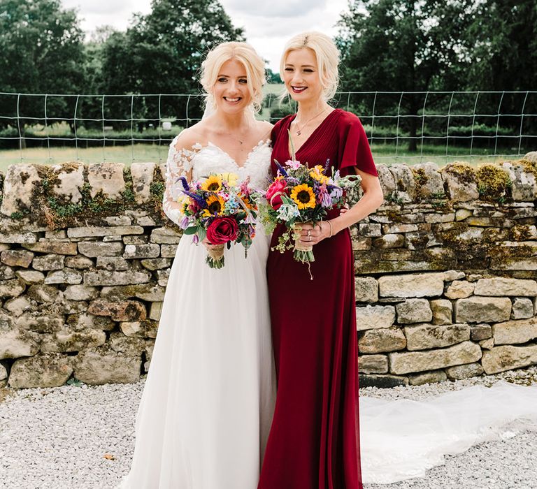 Bride in Pronovias wedding dress stands beside her bridesmaid in red bridesmaid dress complete with silk waistband and short sleeves