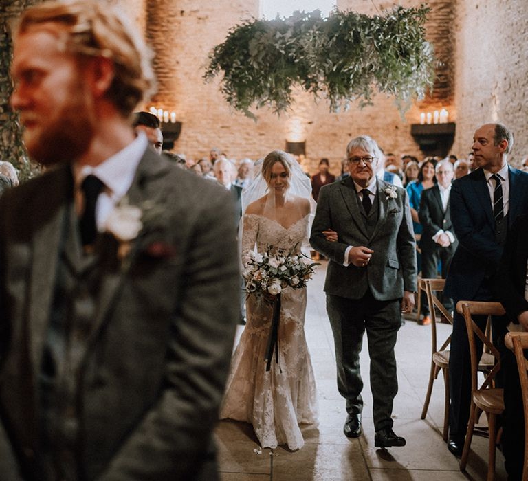 Father of the bride in a grey woollen suit walks the bride in her lace mermaid wedding dress down the aisle to the groom at the end 