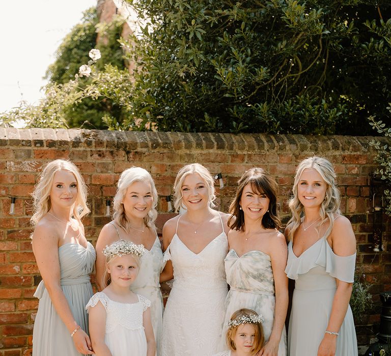 Bridesmaids wear different styled sage green dresses whilst stood beside bride in lace fitted wedding dress