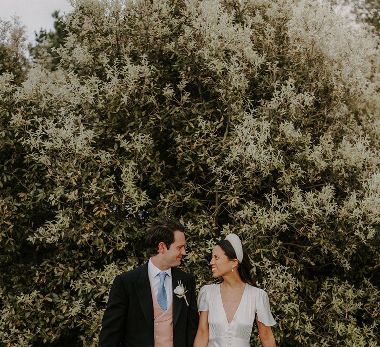 Groom in black morning suit with pinstriped trousers, pink waistcoat and blue tie with bride in Ghost satin wedding dress and pearl headband