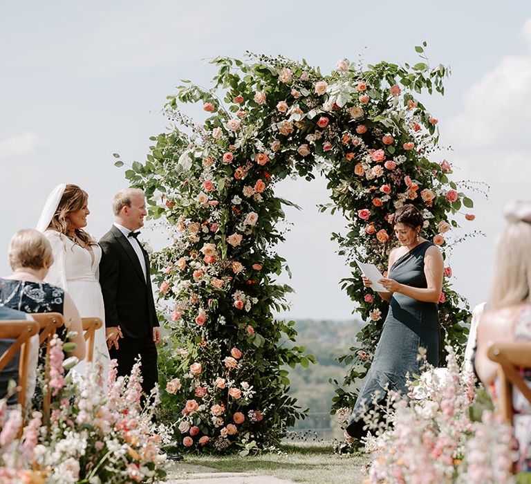 Bridesmaid in one shoulder green bridesmaid dress reads in front of floral archway filled with orange and pink roses surrounded by green foliage