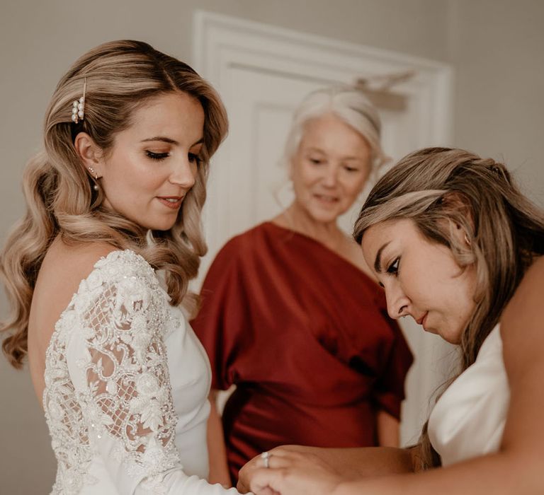 Bridesmaids in champagne gold helps the bride into her wedding dress with waved hair and pearl hair clips 