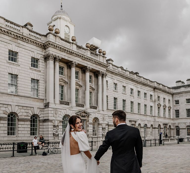 Bride in open back detail wedding dress with long sheer veil and groom in dark tux holding hands and walking around Somerset House wedding venue