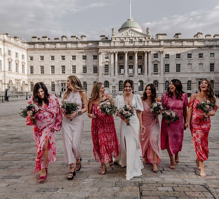 Bride standing with bridal party wearing mismatched floral and plain pink bridesmaid dresses of varying lengths and fabrics