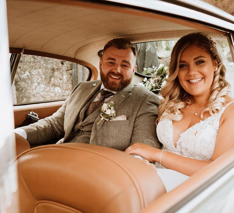 Bride and groom sit in the back of the grey Aston Martin wedding car 