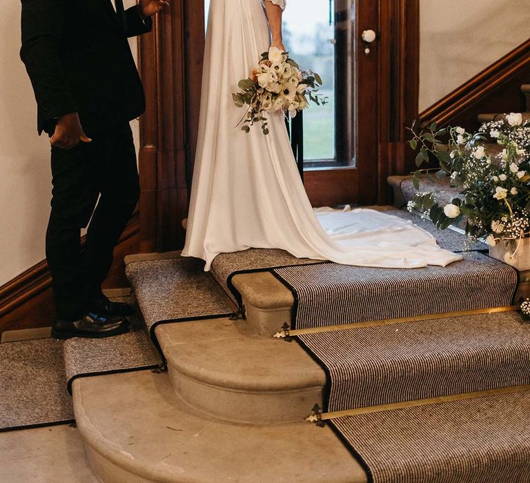 Groom in black tux meeting bride in satin quarter length sleeve wedding gown and minimalist floral bouquet at the top of the stairs