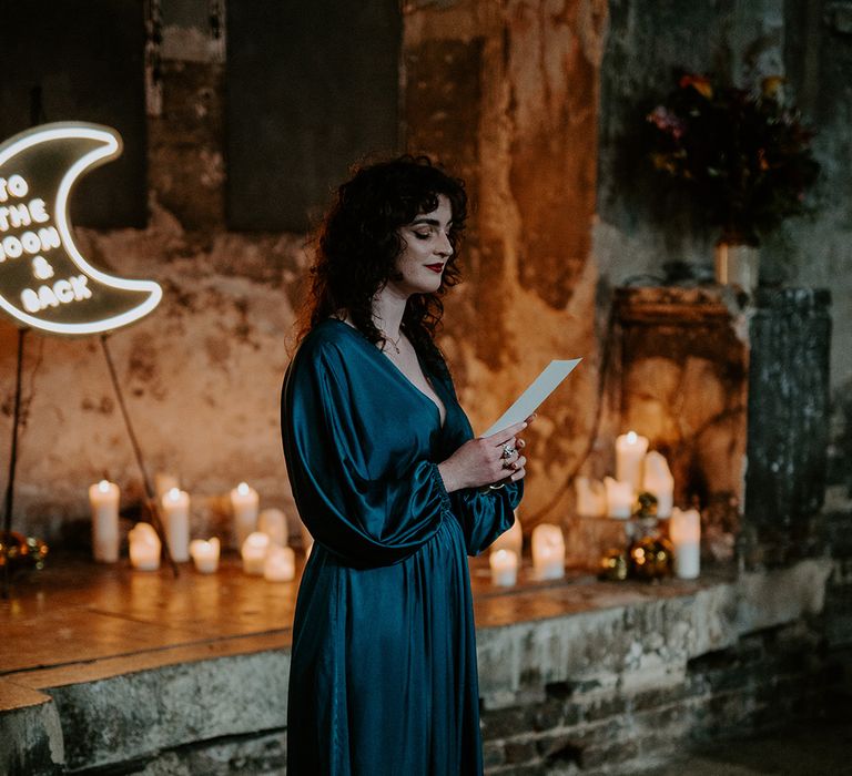 Bridesmaid in satin green dress doing a wedding reading at The Asylum Chapel surrounded by candles and a moon neon sign