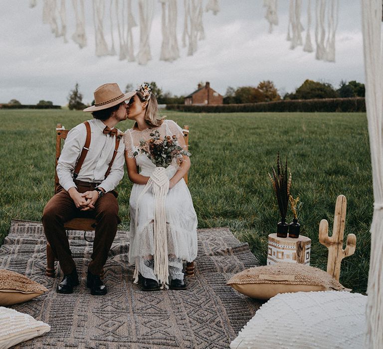 The bride and groom sit down on wooden chairs and share a kiss at their outdoor boho set up 