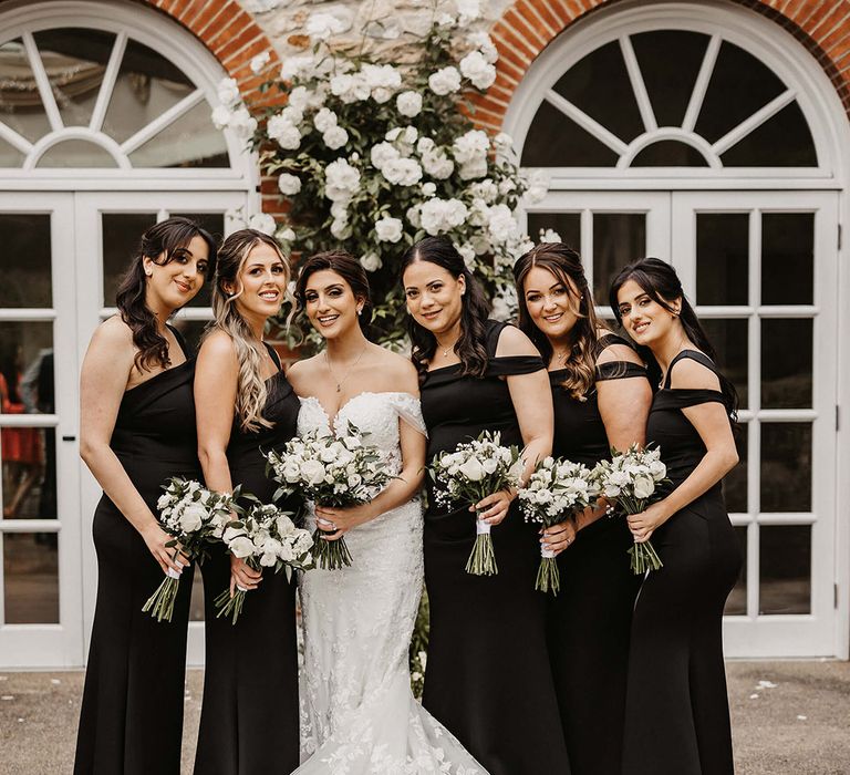 Bride in a flower lace wedding dress and bridesmaids in one shoulder black dresses holding white flower bouquets 