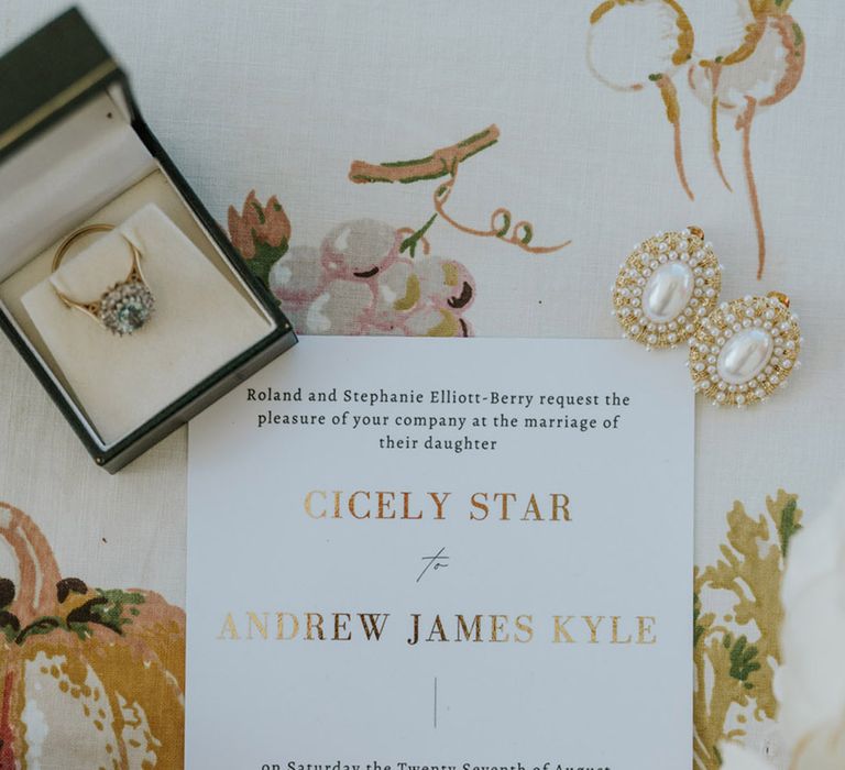 Black and white wedding invitation with gold foil surrounded by the bride's engagement ring with gold earrings 