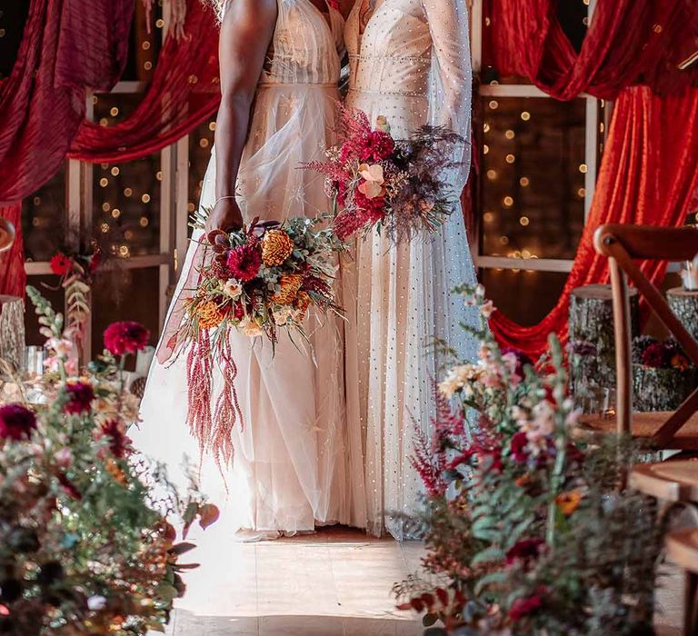 Bride in crystal embellished gown and gold headdress and sleeveless floaty peach gown and gold leaf crown standing with red wedding drapes, fairy lights and autumnal dried flower arrangements